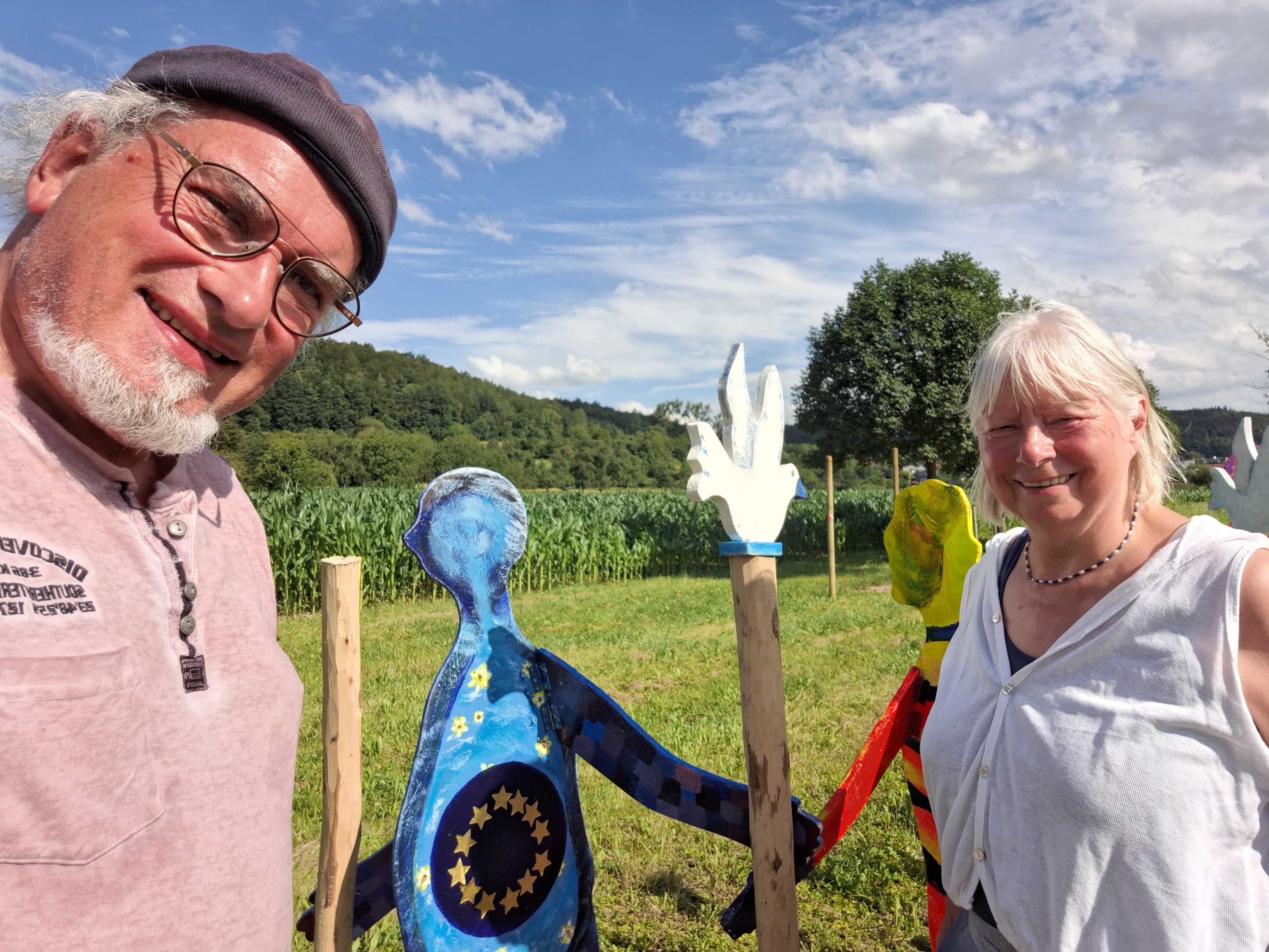 Alexander und Regina stehen lachend auf einer Wiese neben zwei bemalten Holzfiguren, im Hintergrund ein Maisfeld und bewaldete Hügel