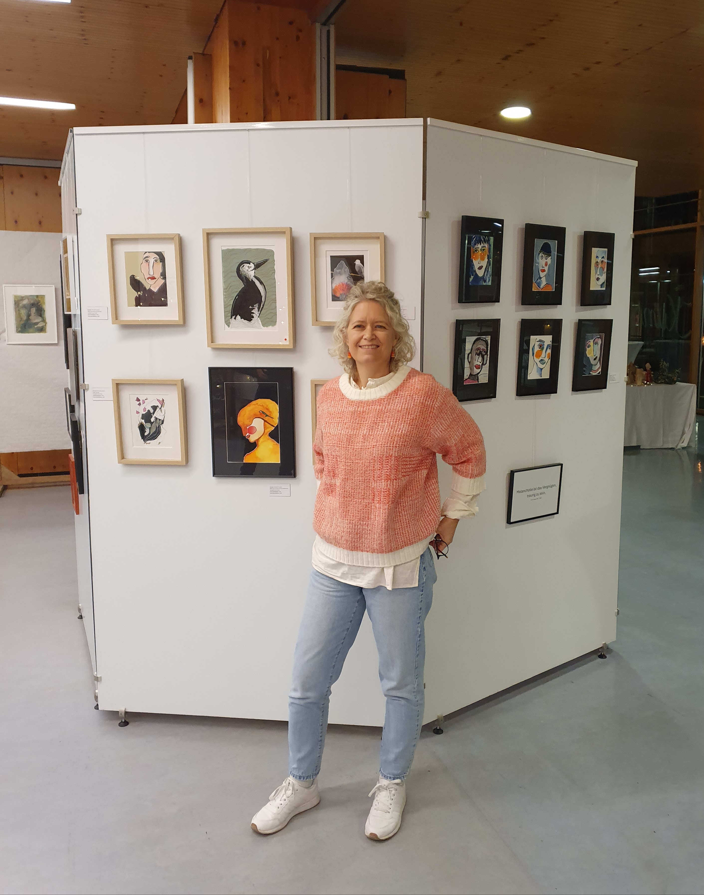 Elisabeth le Blanc steht lächelnd vor einer Ausstellungswand mit gerahmten Porträtzeichnungen