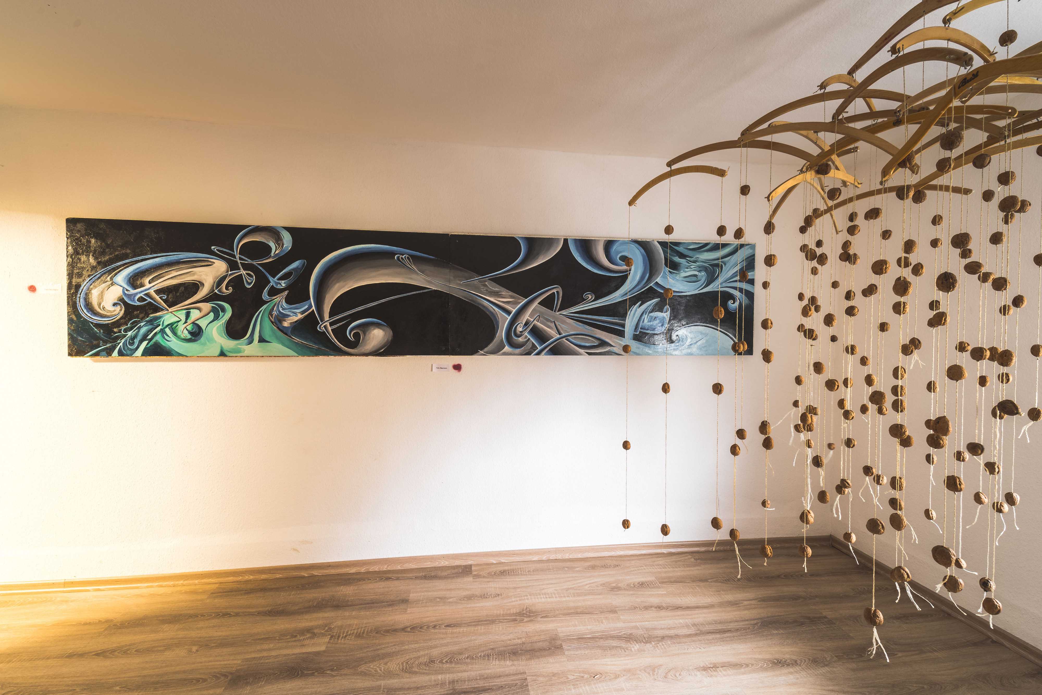 Ausstellungsraum mit einem länglichen abstrakten Wandbild in Blau, Grau und Grün sowie einer Installation aus an Schnüren hängenden Walnüssen