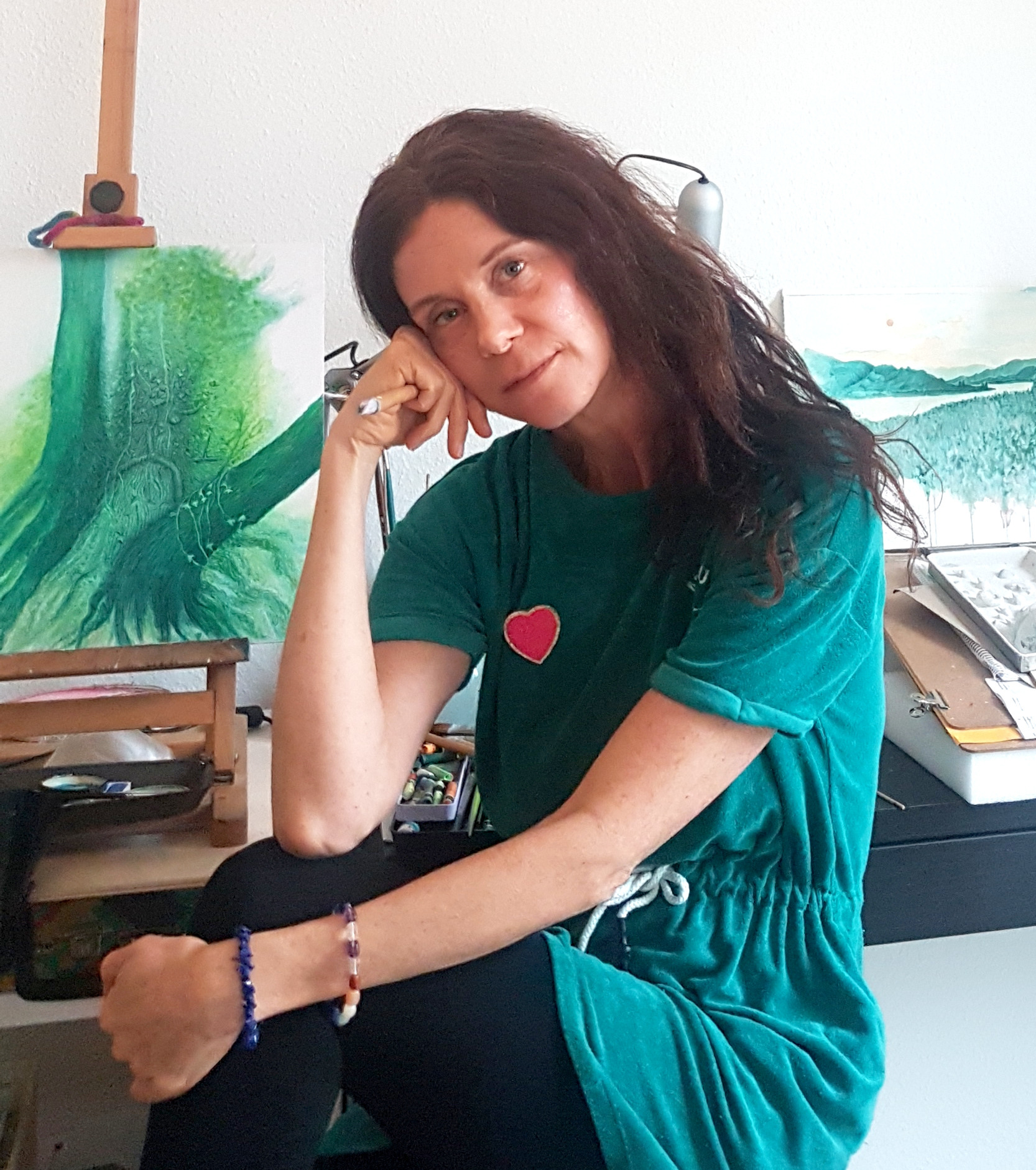 Ines Vetter sitzt nachdenklich in ihrem Atelier, im Hintergrund grüne Landschaftsgemälde auf Staffeleien