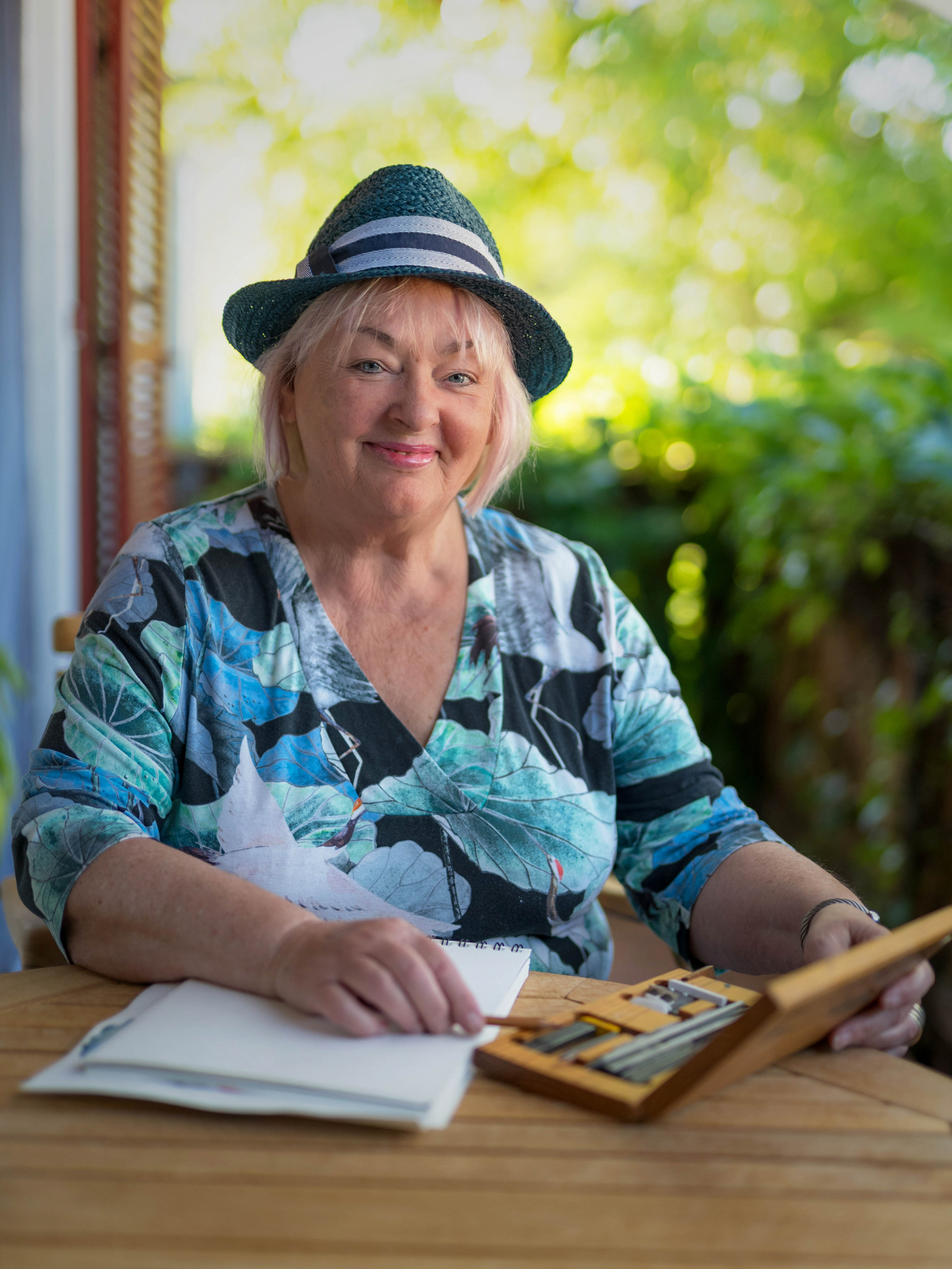 Karola Kuhn mit Hut an einem Tisch sitzend, ein Skizzenbuch vor sich