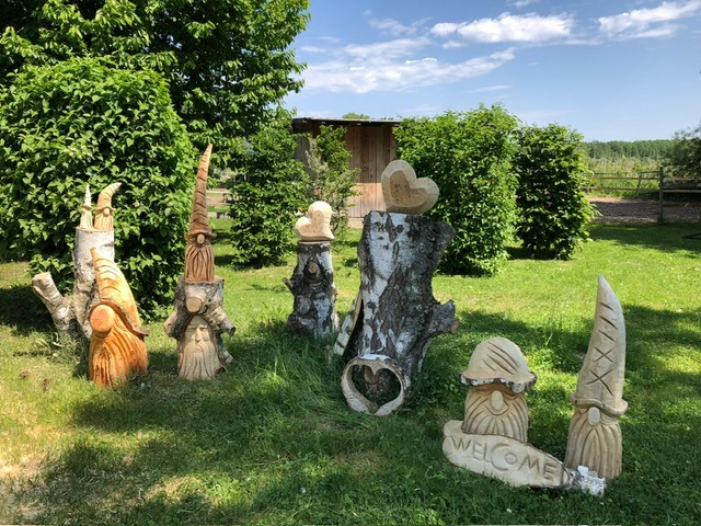 Gruppe geschnitzter Wichtel- und Eulenfiguren aus Holz auf einer Wiese, dazwischen ein Schild mit der Aufschrift WELCOME