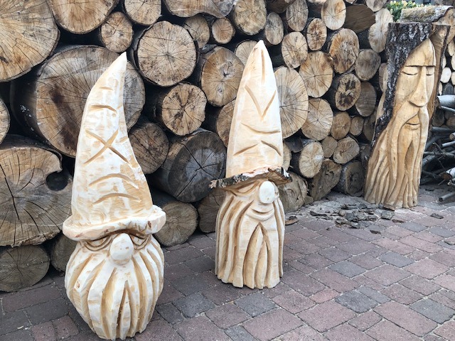 Zwei geschnitzte Wichtelfiguren und ein Gesicht in einem Baumstamm vor einem Holzstapel