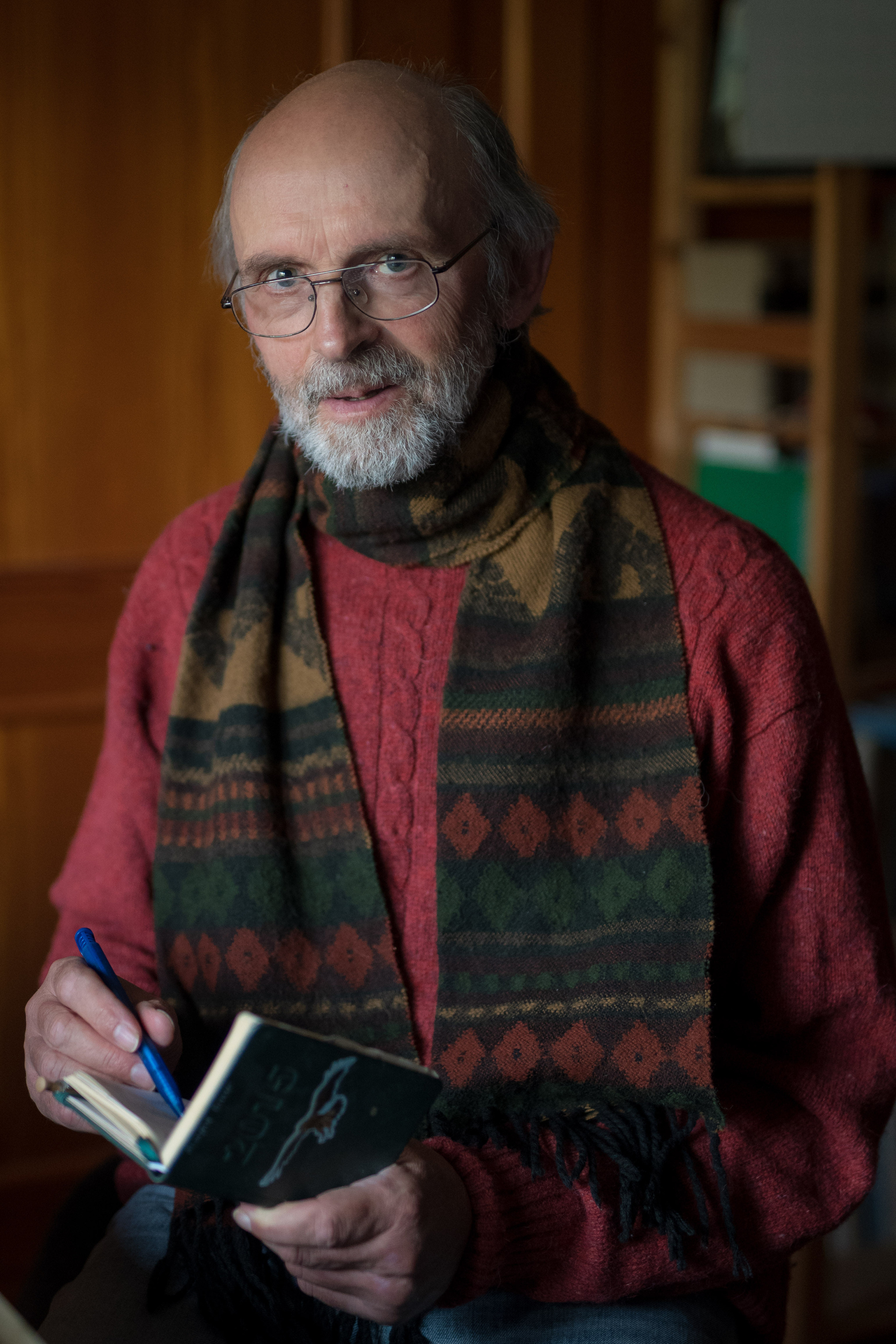 Porträtfoto von Dr. Manfred Lieser, ein älterer Mann mit Brille und weißem Bart, in rotem Pullover und gemusterten Schal, schreibt mit blauem Kugelschreiber in ein kleines Notizbuch