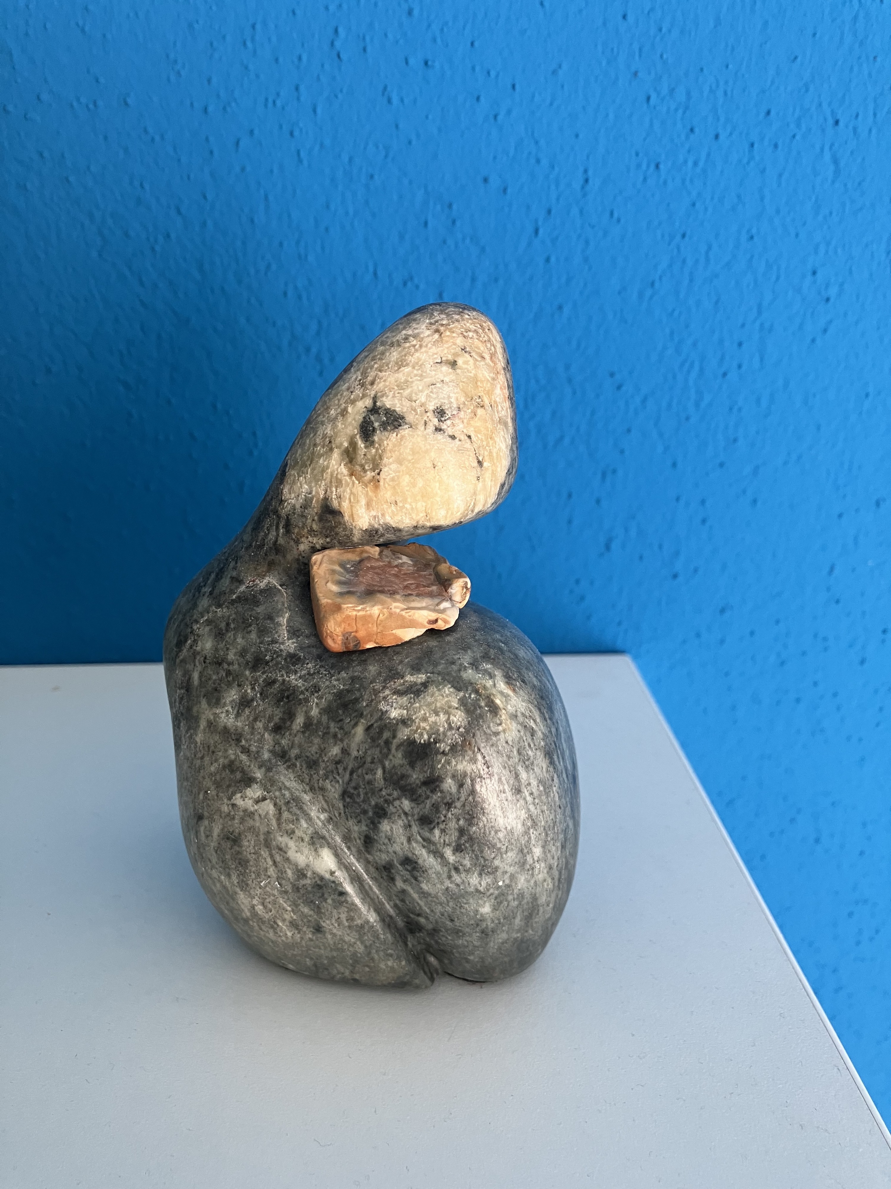 Speckstein-Skulptur in Form eines geneigten Kopfes, graues Gestein mit kleinem hellem Einsatz, vor blauem Hintergrund