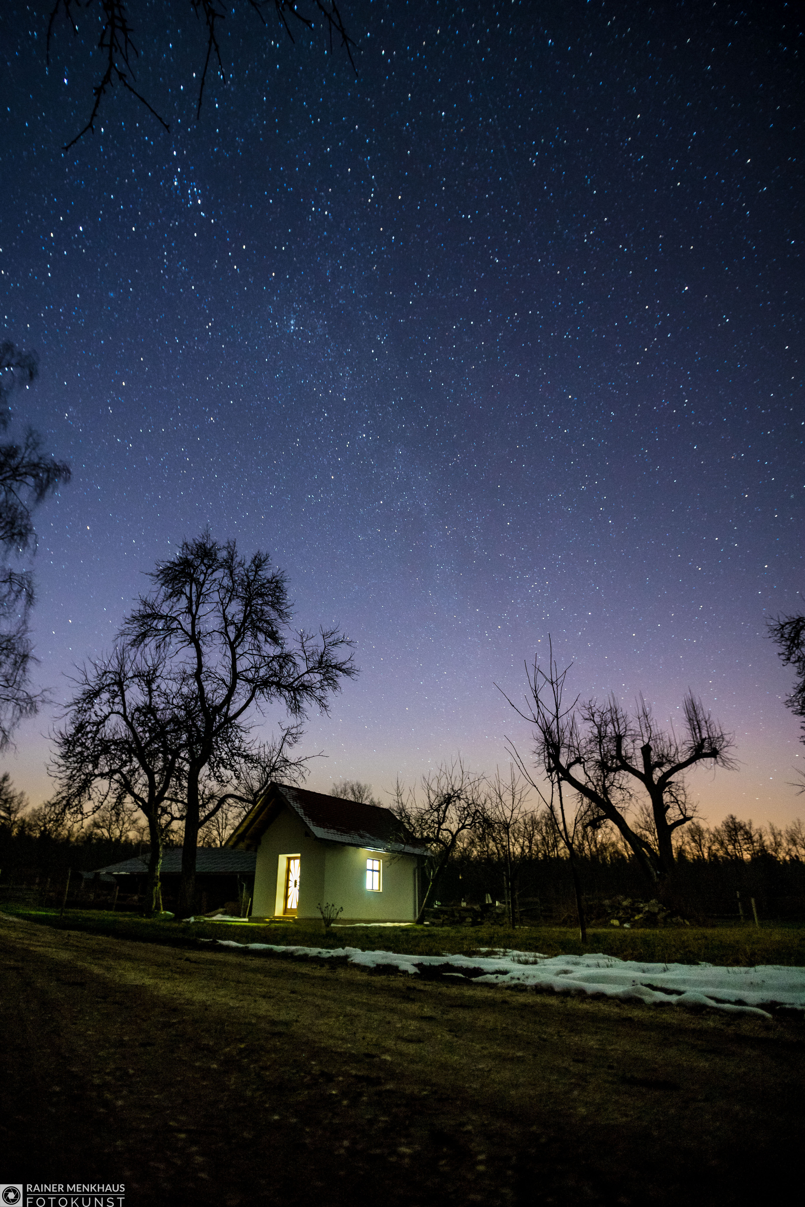 Sternenhimmel über einem beleuchteten kleinen Haus zwischen kahlen Bäumen bei Nacht