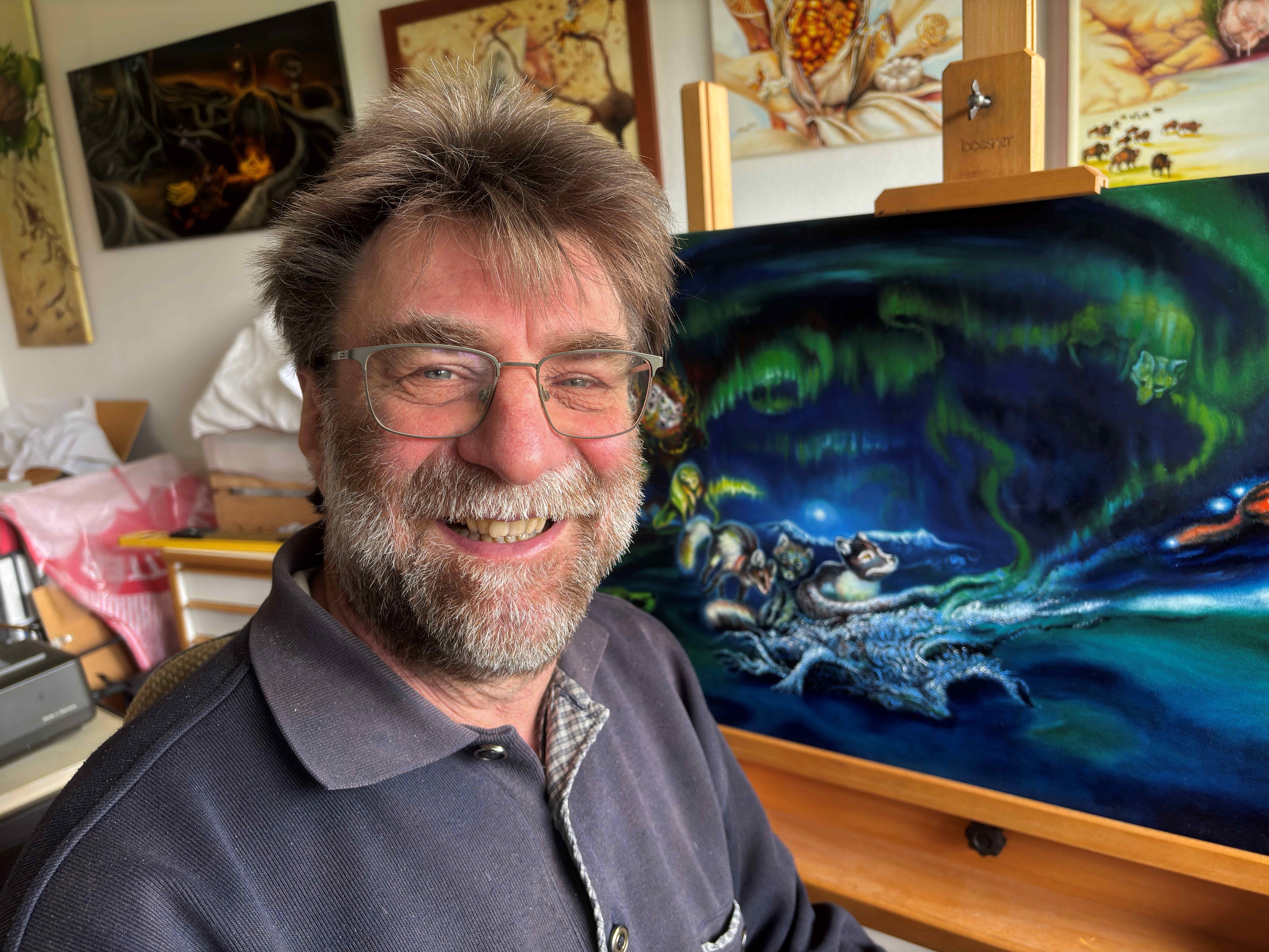 Porträtfoto von Roland Spohn, lächelnd mit Brille und grau meliertem Bart, im Atelier vor gerahmten Gemälden und einer Staffelei mit einem Nachtbild von Tieren