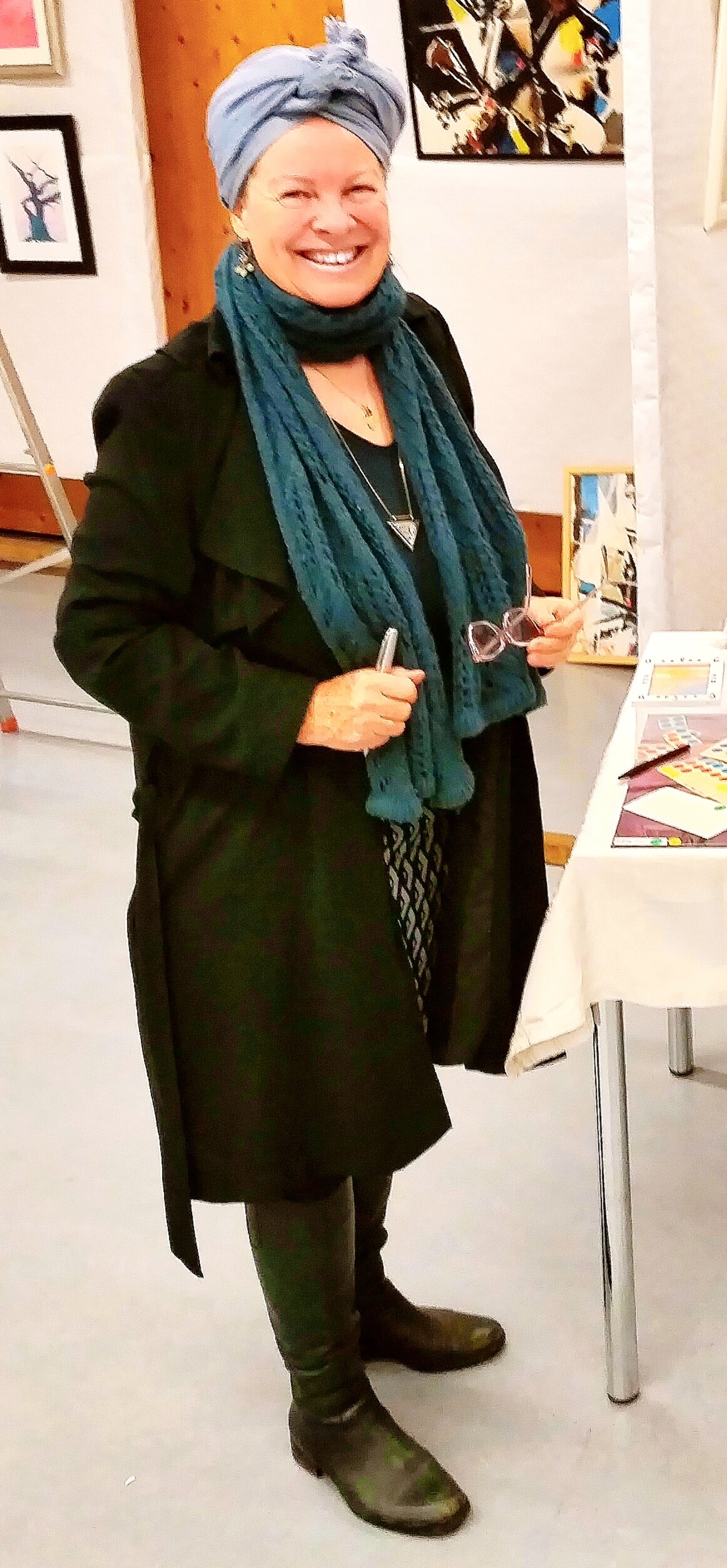 Safina Aksoy lächelt bei einer Ausstellung, mit blauem Kopftuch und türkisfarbenem Schal, hinter ihr gerahmte Bilder
