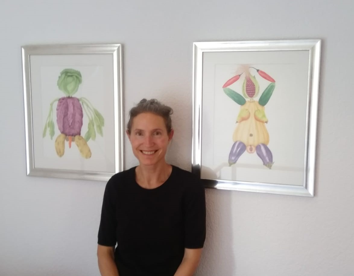 Vanessa Pasqualetto lächelt vor zwei gerahmten botanischen Aquarellen, die Figuren aus Gemüse und Früchten zeigen