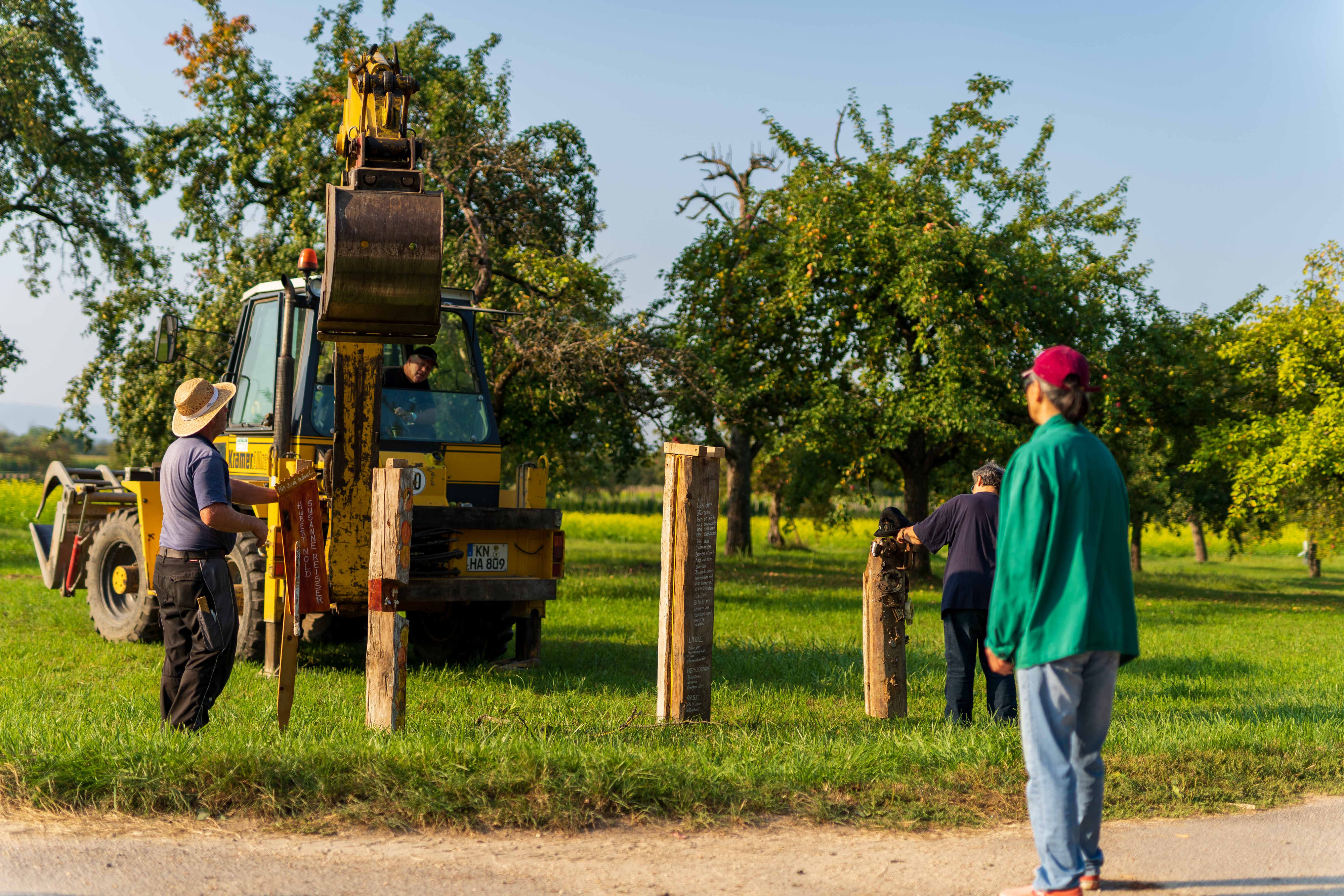 Drei Personen stellen mit einem Bagger bemalte Holzpfosten in einer Streuobstwiese auf