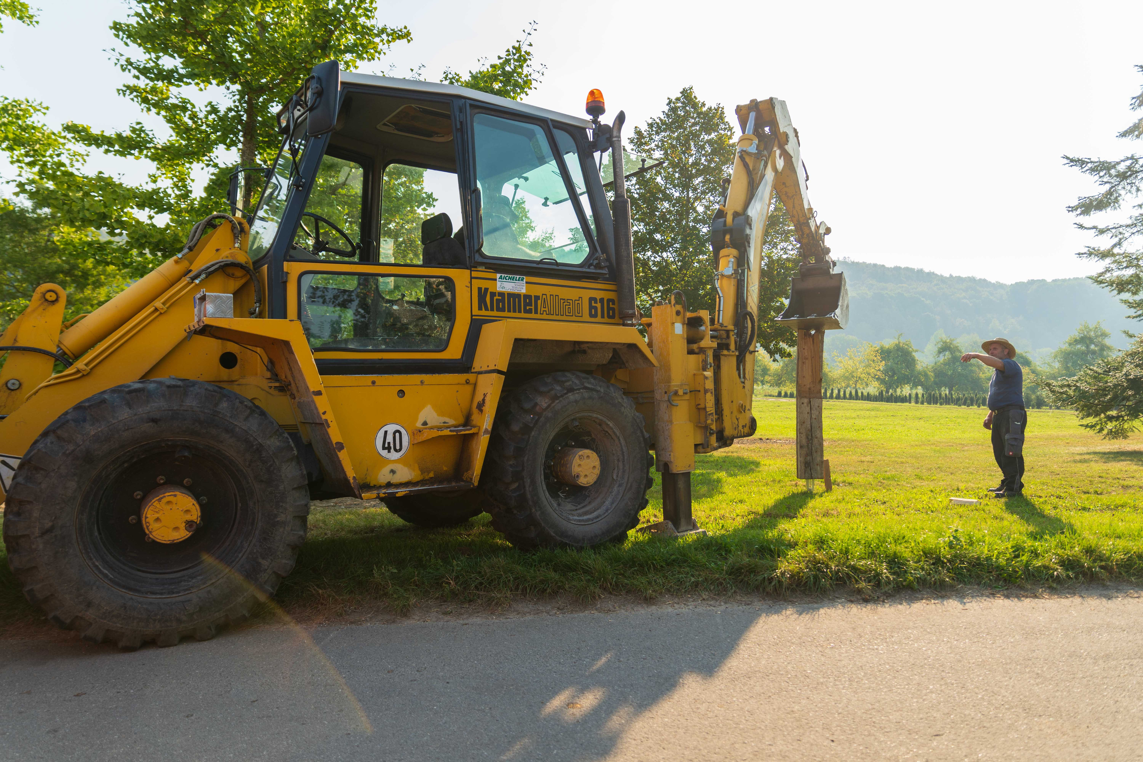 Bagger setzt einen Holzpfosten am Feldweg, ein Helfer steht daneben