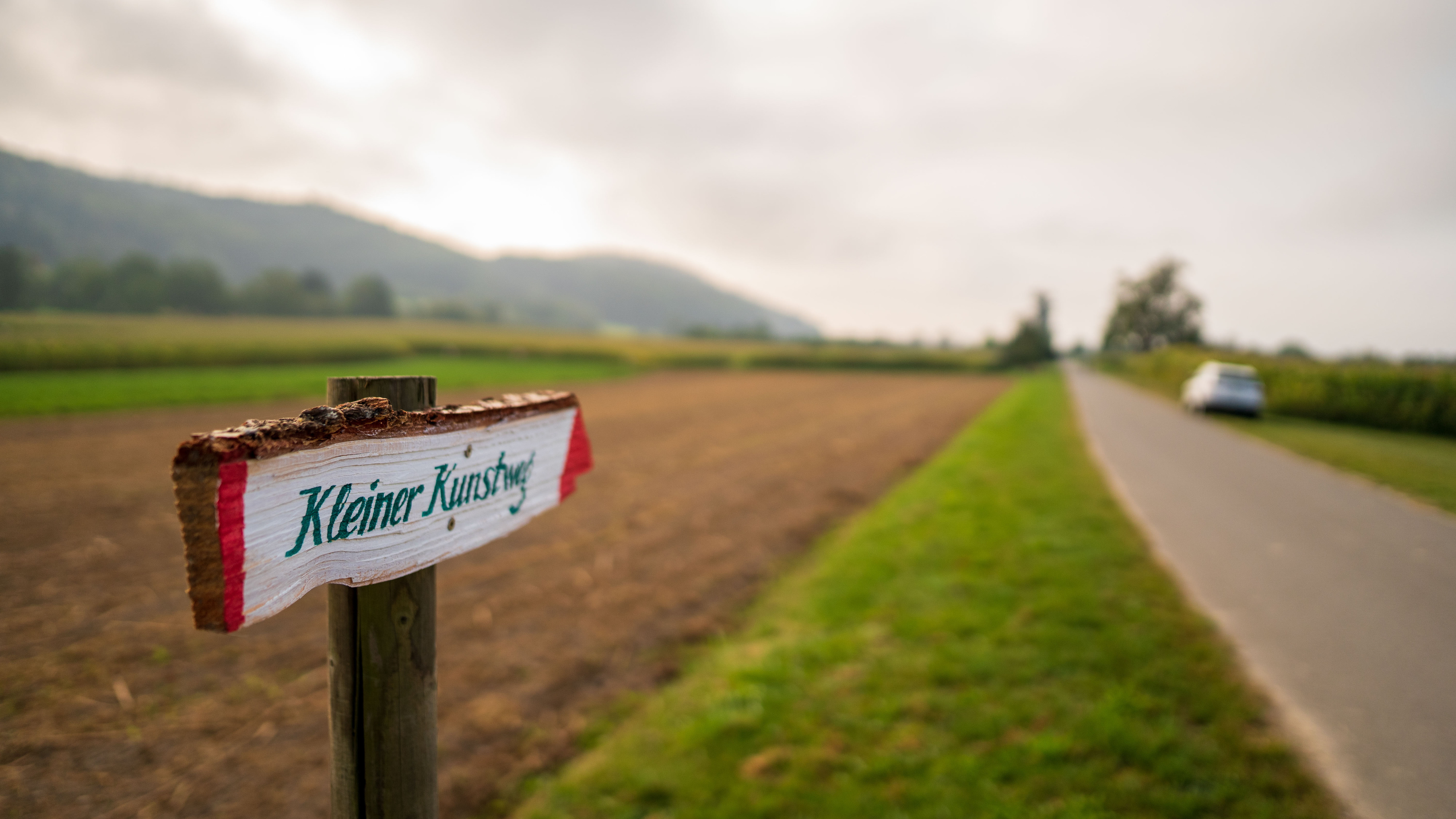 Hinweisschild „Kleiner Kunstweg" an einem Holzpfosten, dahinter ein Feldweg zwischen Wiesen