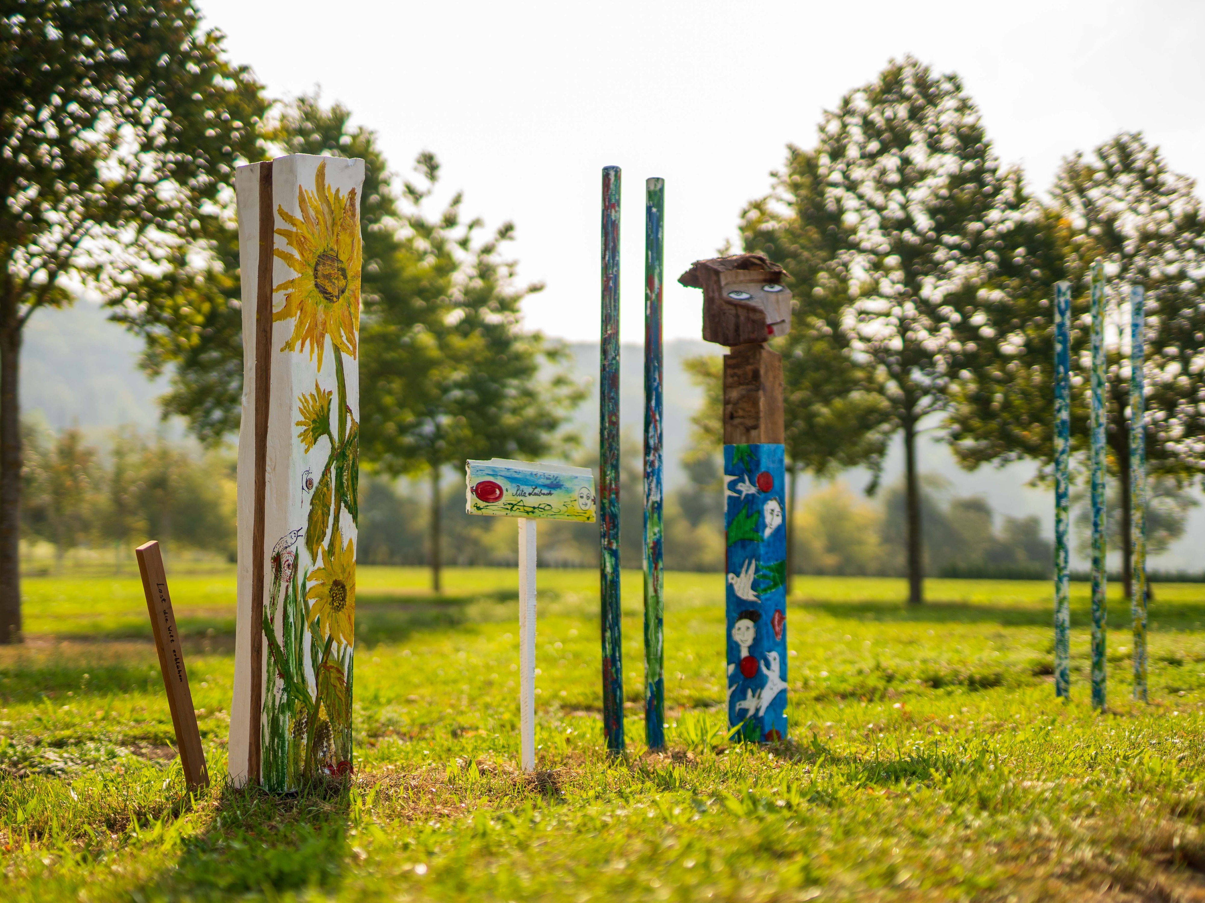 Bemalter Pfosten mit Sonnenblumenmotiv neben weiteren Kunstpfosten am Wegrand
