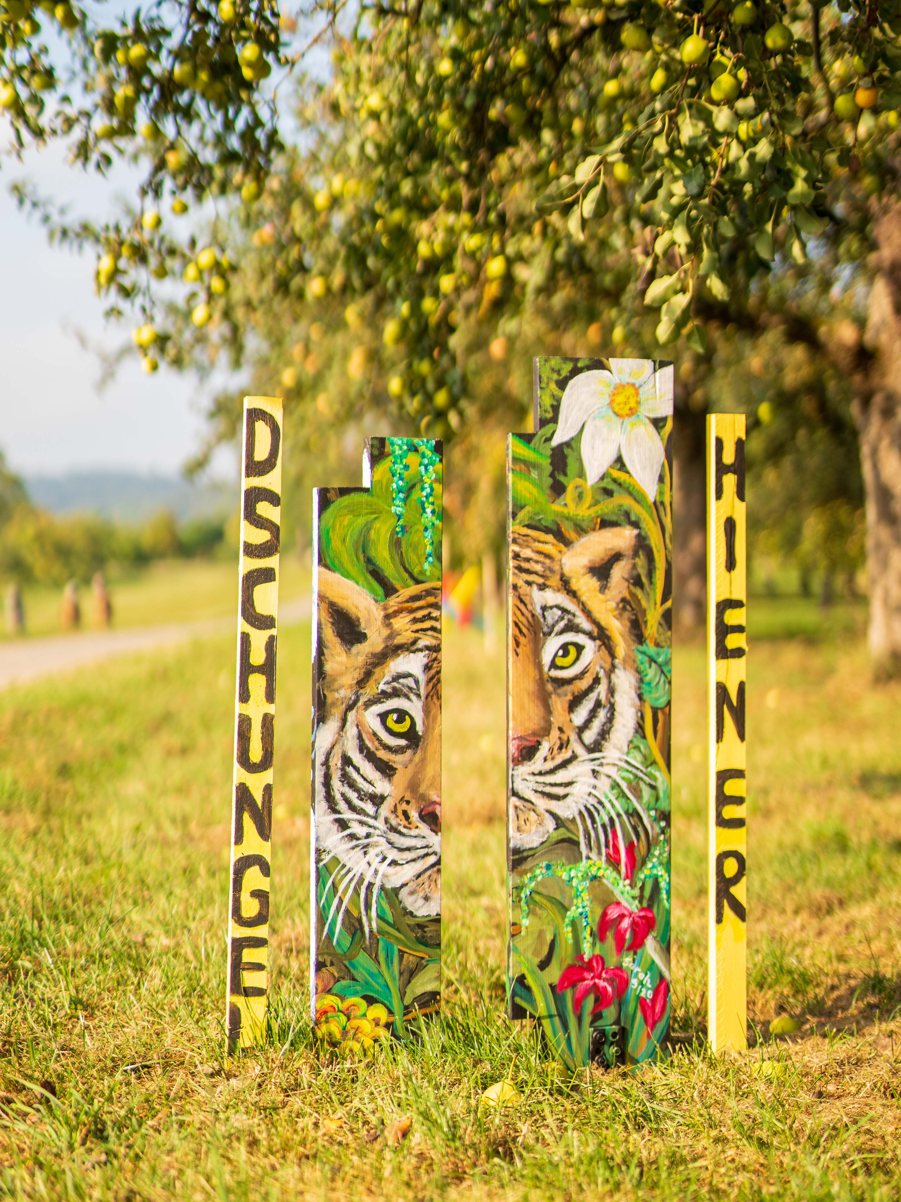 Zwei schmale Holzpfosten mit den Wörtern „Dschungel" und einem gemalten Tiger sowie Blüten, zwischen Ästen platziert