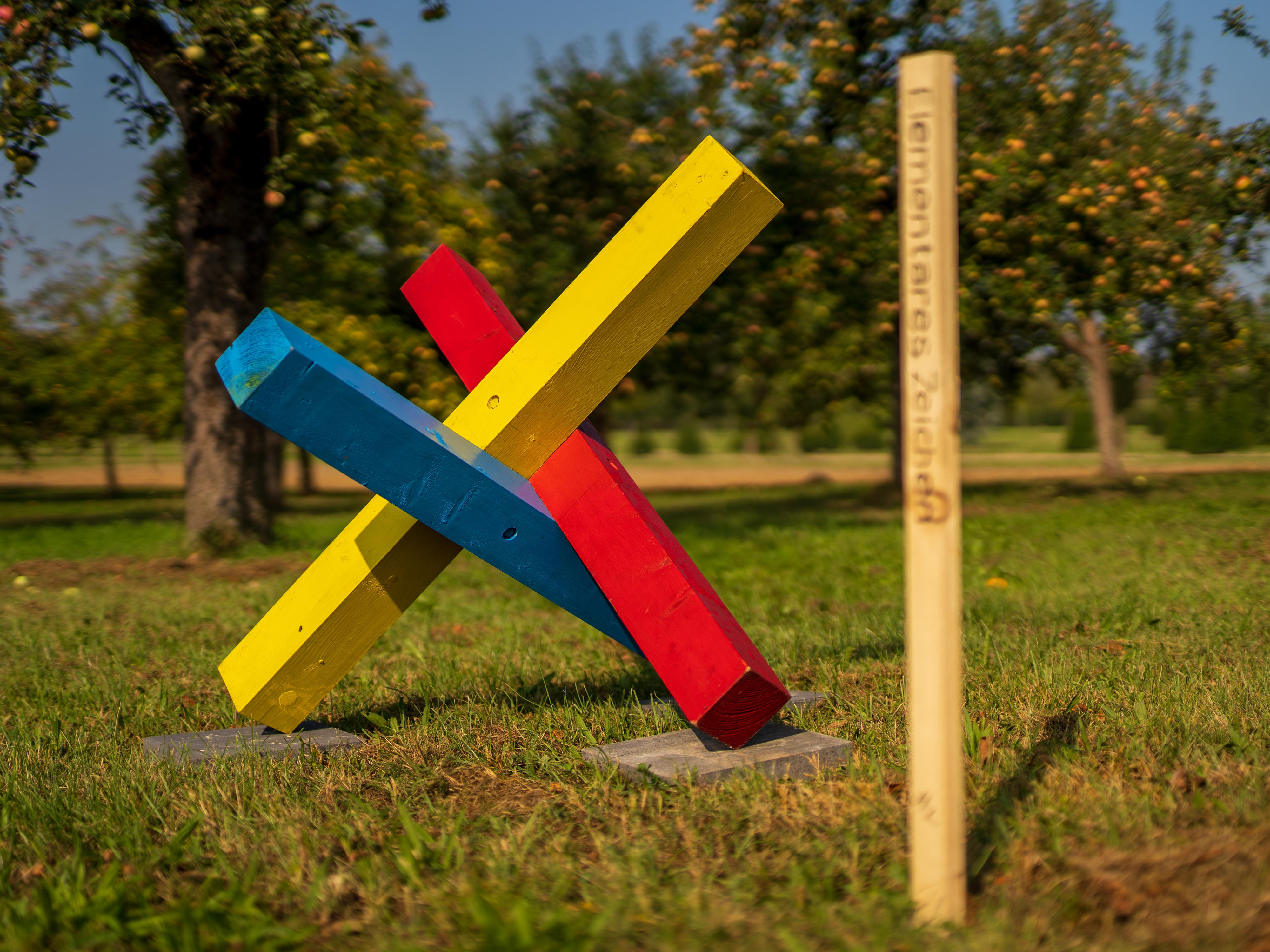 Skulptur aus gekreuzten, farbig bemalten Holzlatten in Rot, Gelb und Blau neben einem schlichten Pfosten auf einer Wiese