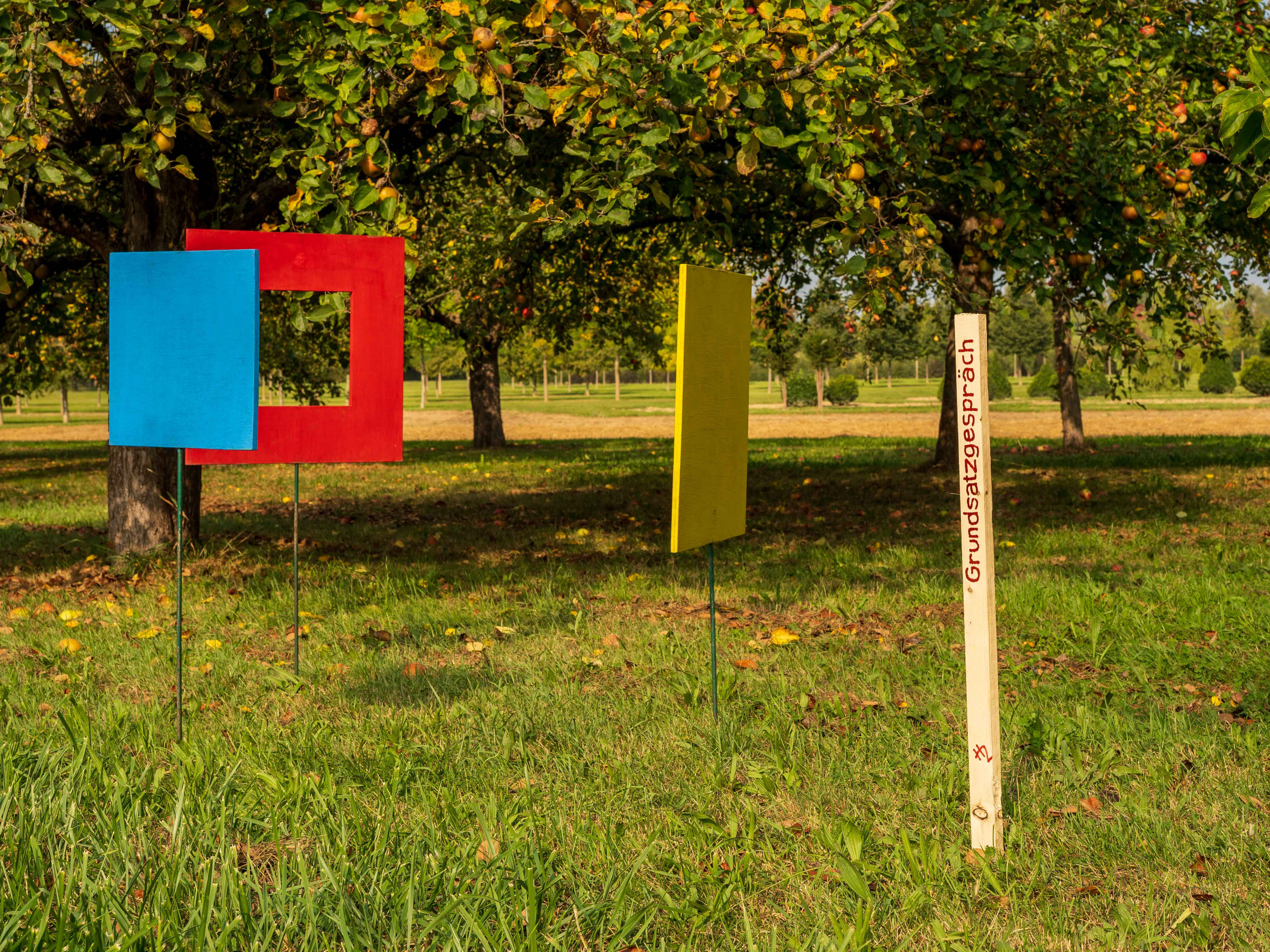 Installation aus einem roten und einem blauen geometrischen Rahmen sowie einem gelben Element auf Pfosten, im Hintergrund Obstbäume