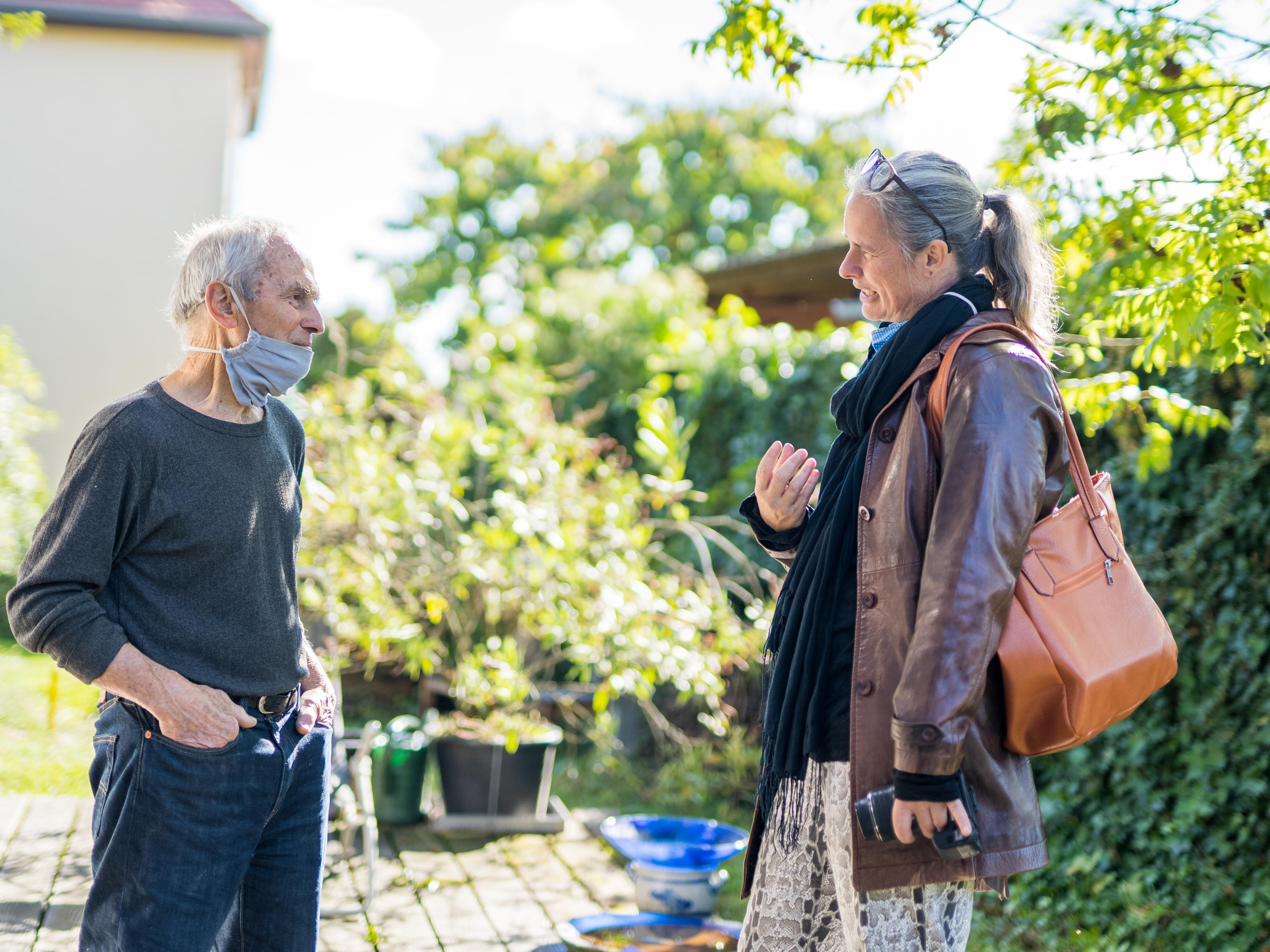 Zwei Personen mit Mund-Nasen-Schutz unterhalten sich im Garten vor einem Atelier