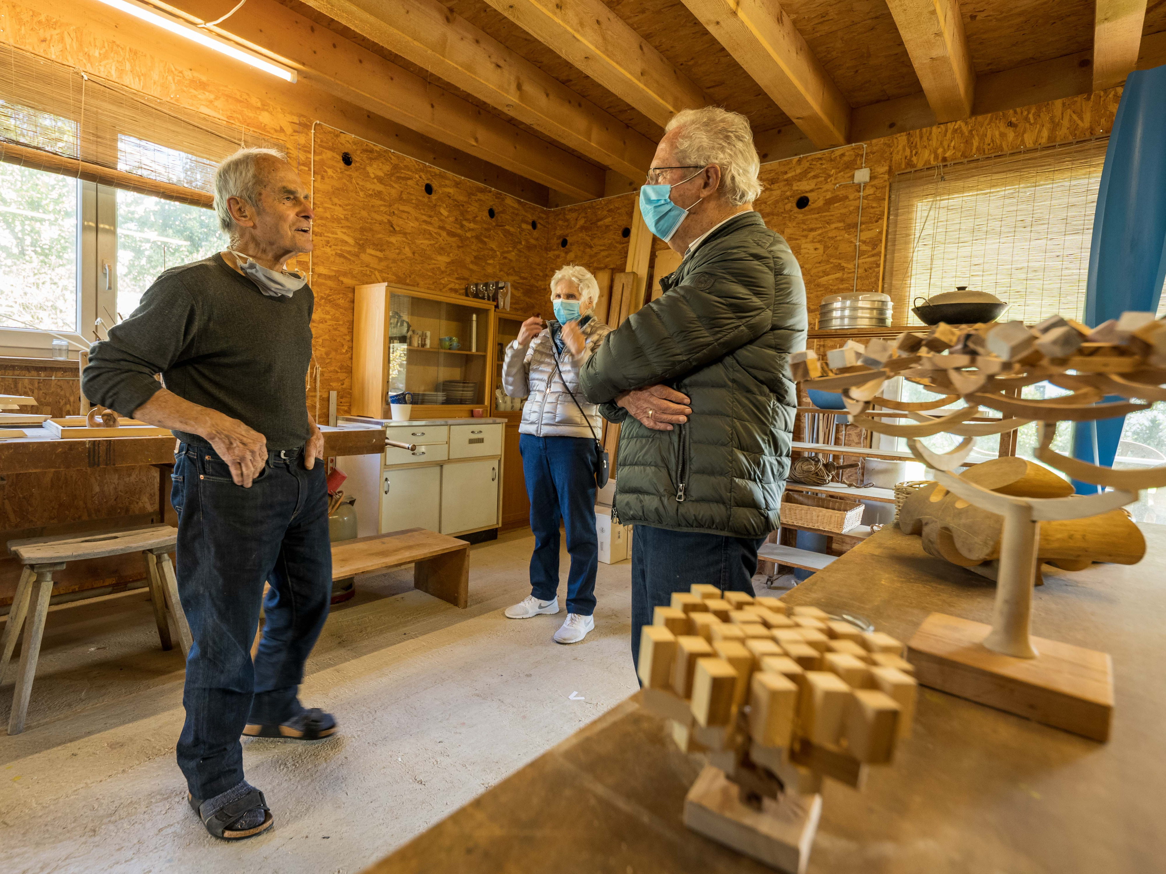 Der Künstler zeigt zwei Besucherinnen mit Mund-Nasen-Schutz sein Atelier mit gedrechselten Holzobjekten auf Werkbänken