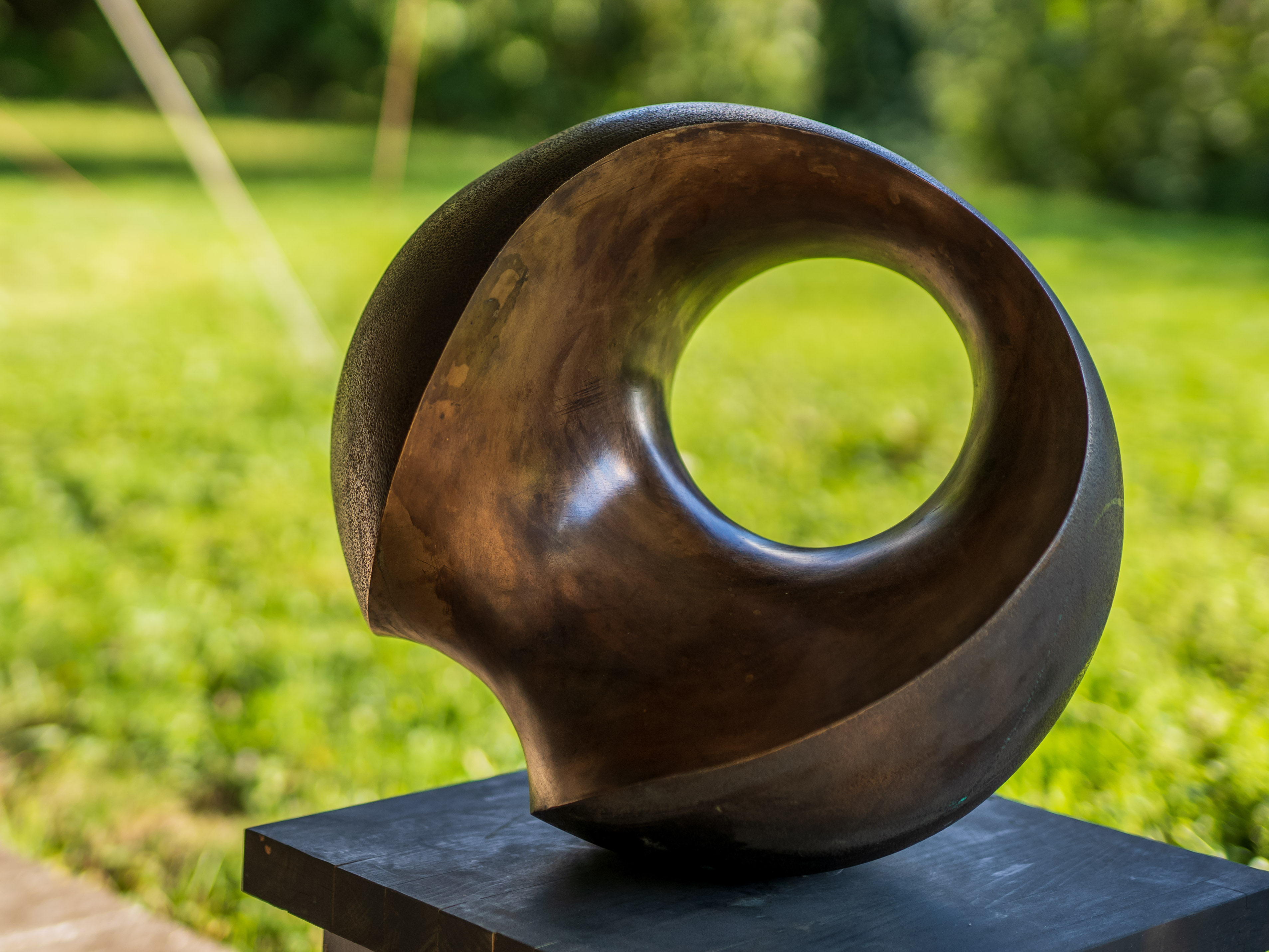Bronzefarbene Skulptur mit kreisförmiger Öffnung auf einem schwarzen Sockel im Garten