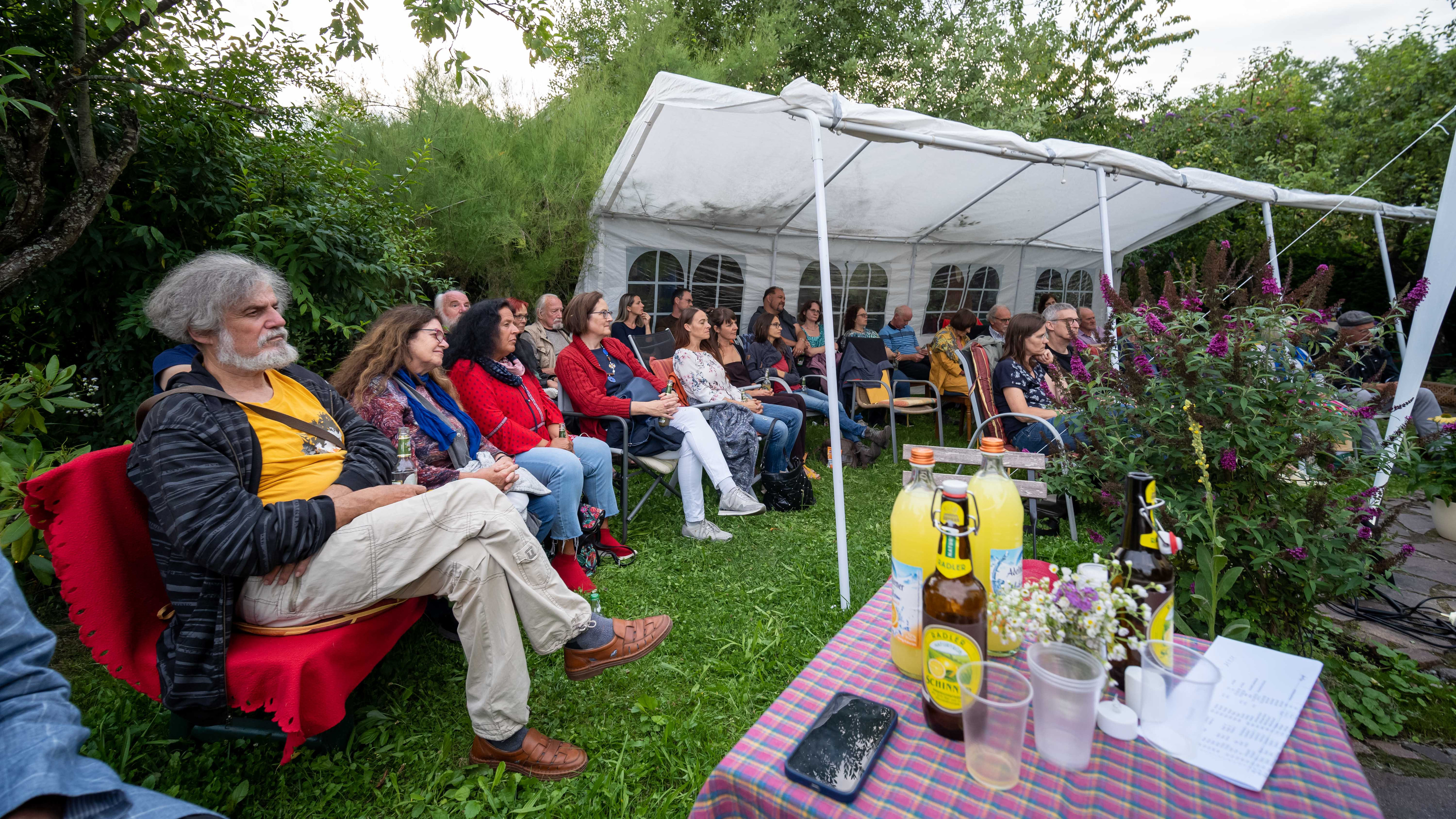 Publikum sitzt unter einem Zeltdach im Garten, im Vordergrund eine Picknickdecke mit Gläsern