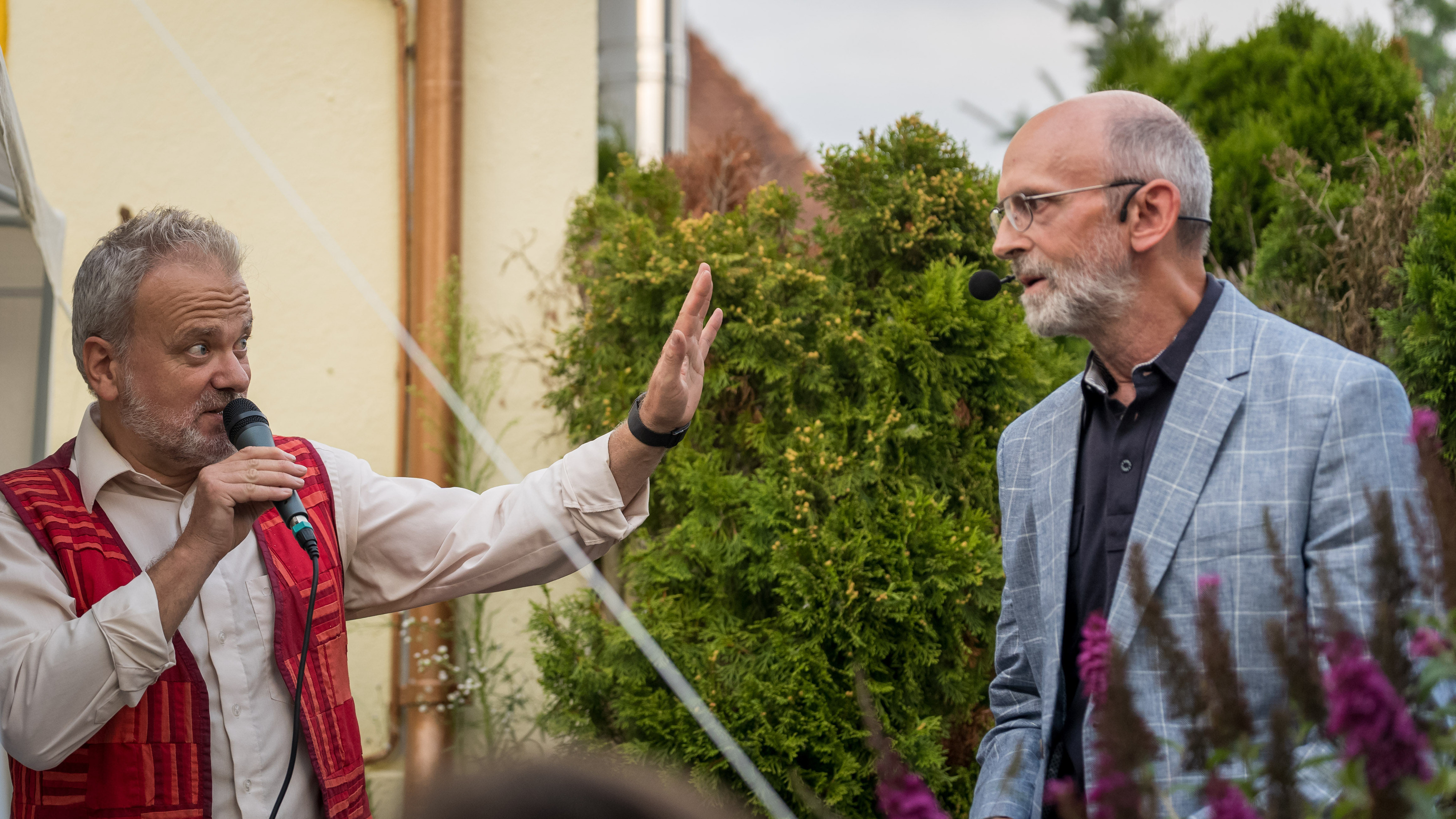 Zwei Männer im Garten, einer in roter Weste gestikuliert im Gespräch