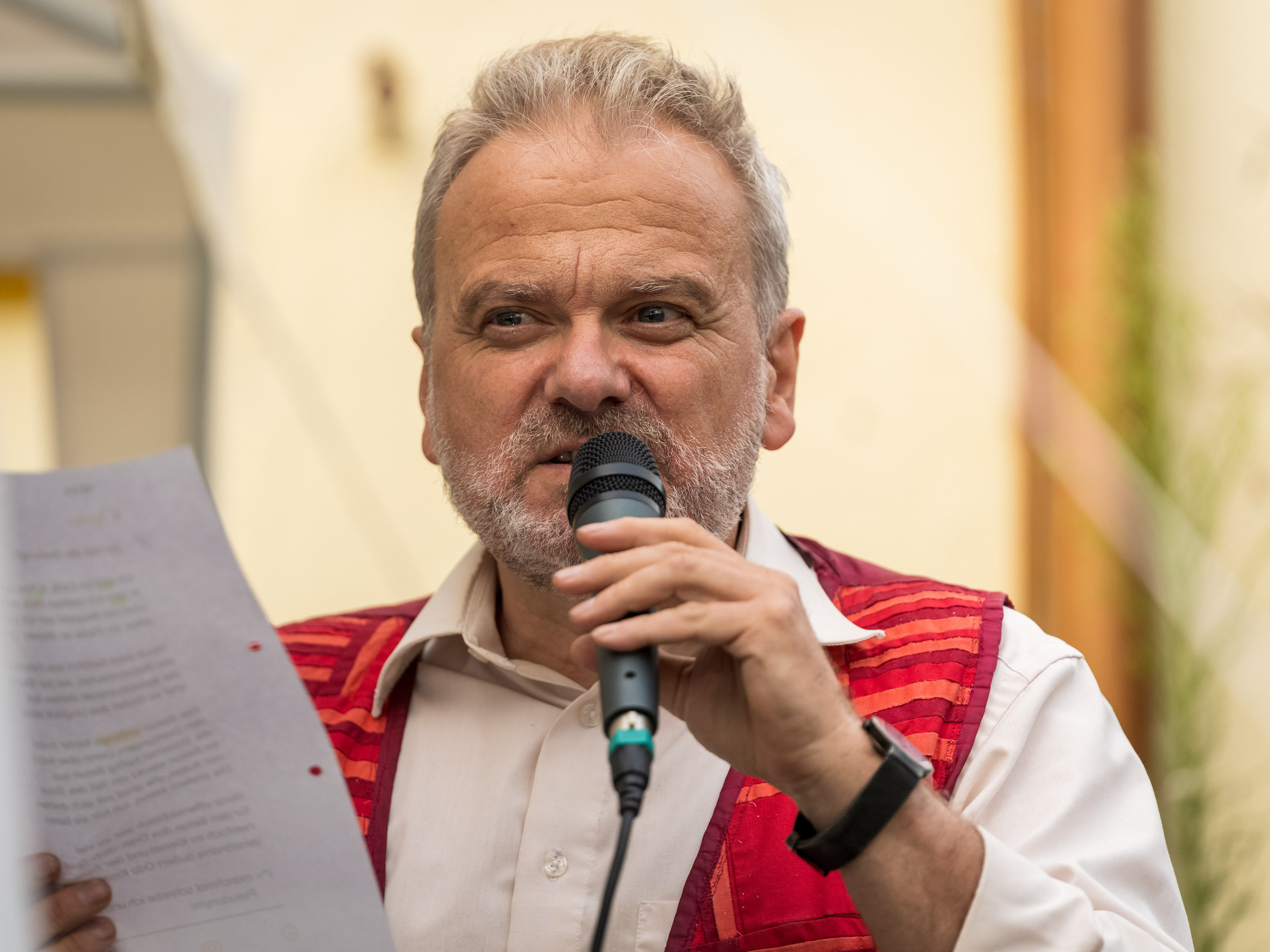 Ein Mann in roter Weste liest mit Mikrofon von einem Blatt Papier ab