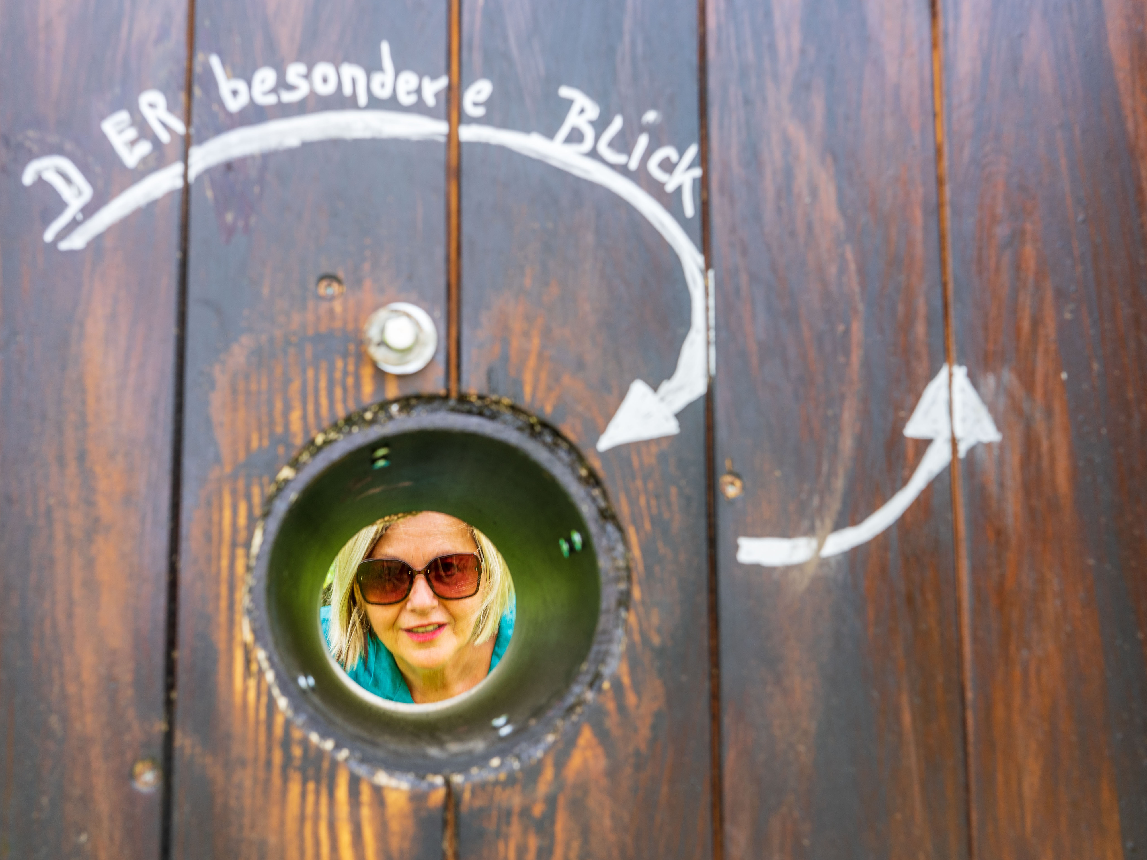 Holzwand mit der Aufschrift „DER besondere Blick" und einem runden Guckloch, durch das das lächelnde Gesicht einer Frau mit Sonnenbrille zu sehen ist