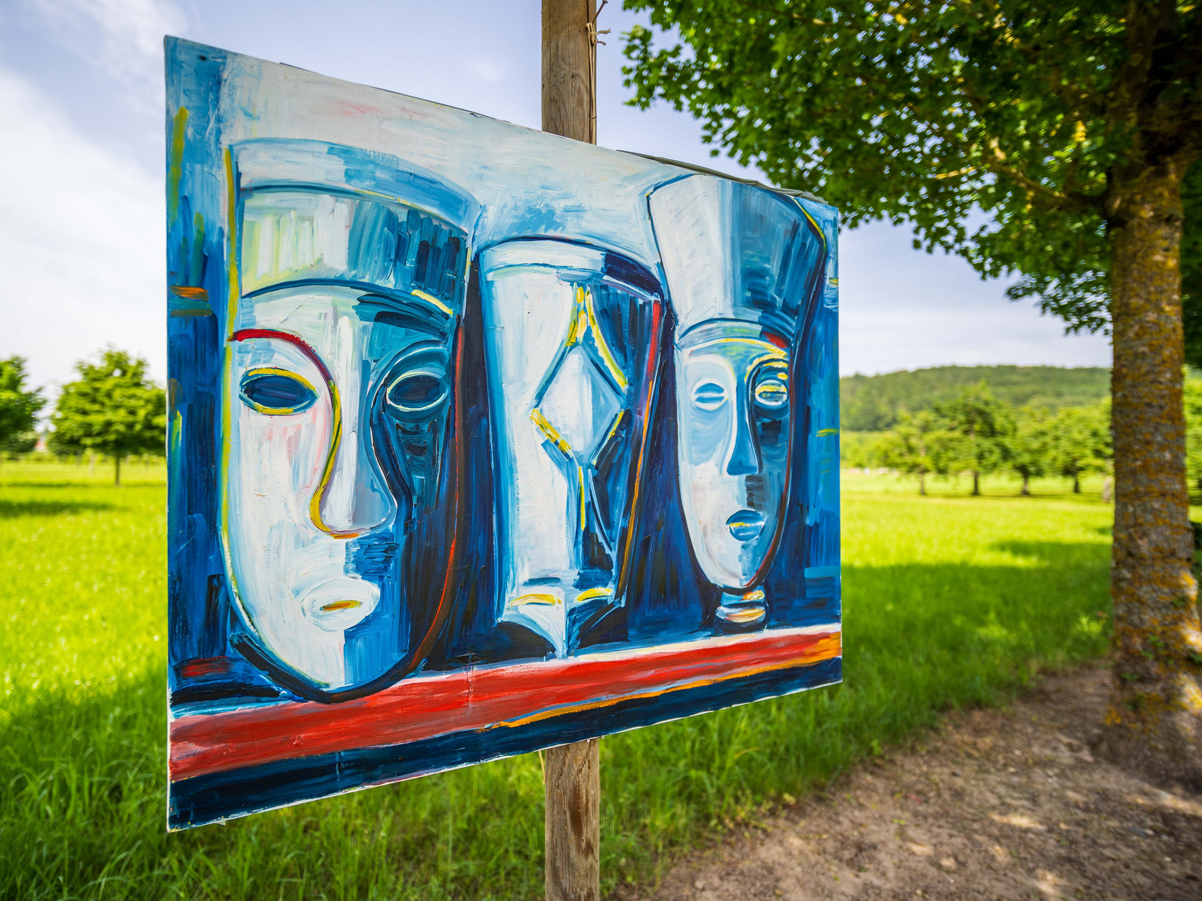 Gemälde mit drei stilisierten blauen Gesichtern, an einem Pfosten in einer Wiese aufgestellt