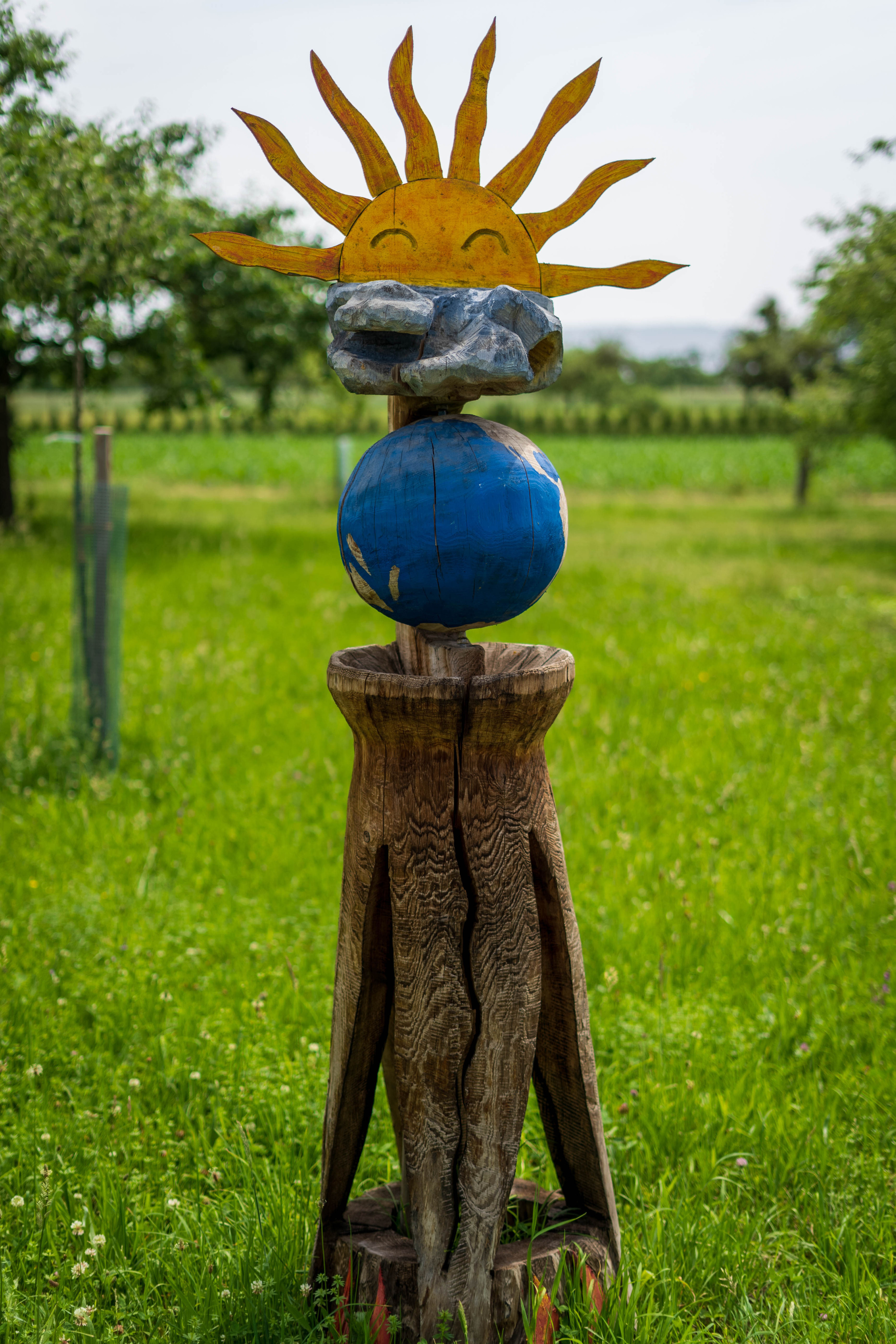Skulptur einer stilisierten Sonne aus Metall auf einer blauen Kugel, auf einem Holzsockel in der Wiese