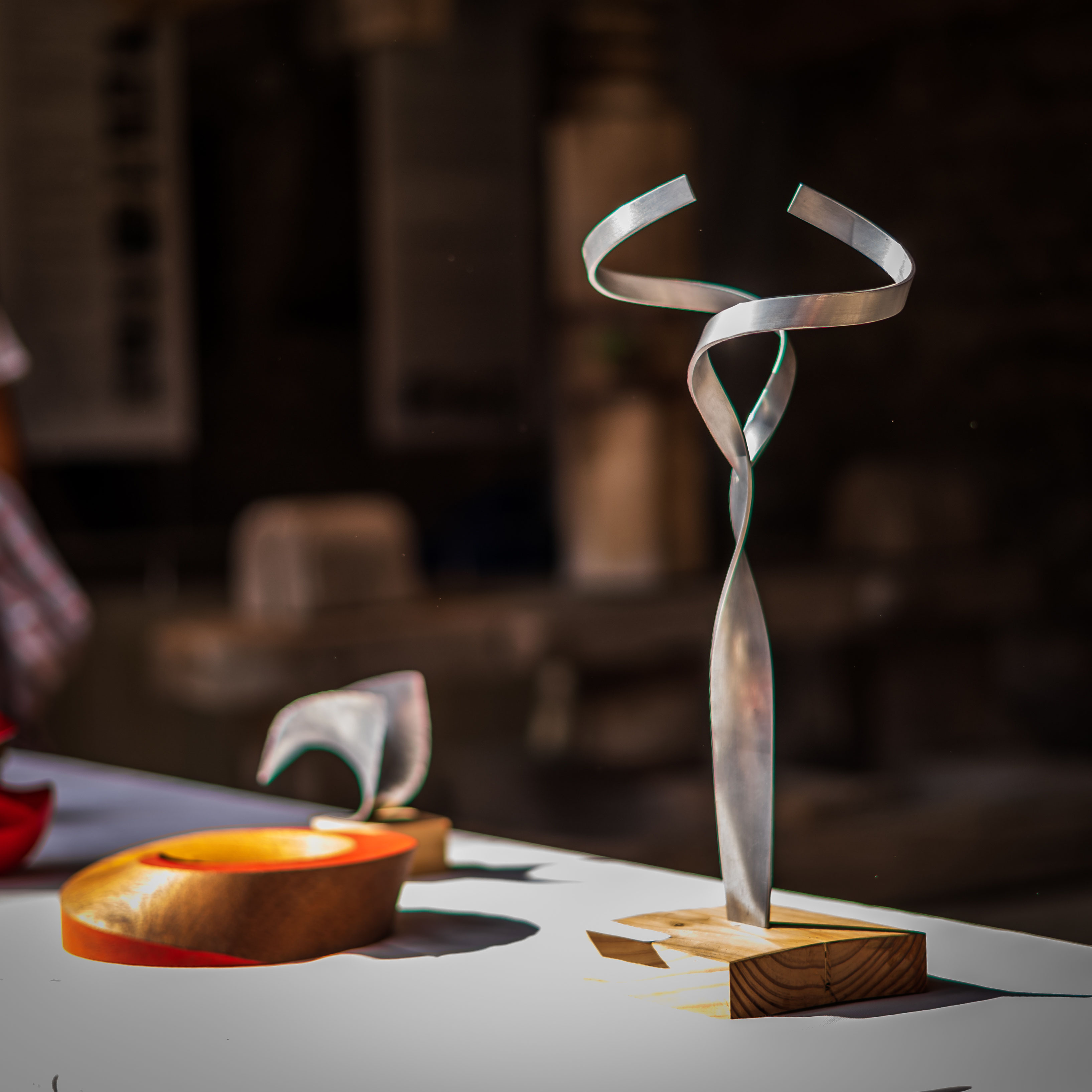 Abstrakte Skulptur aus gebogenem hellem Metallband auf einem Holzsockel, daneben eine Holzschale